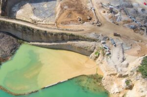 Aerial view of a large quarry with earth-moving equipment. The scene includes excavated land with visible layers, yellow sediment contrasting against green water, and multiple machinery pieces actively involved in extraction and processing.