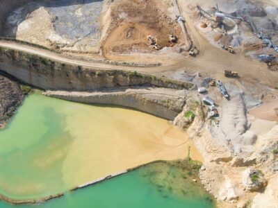 Aerial view of a large quarry with earth-moving equipment. The scene includes excavated land with visible layers, yellow sediment contrasting against green water, and multiple machinery pieces actively involved in extraction and processing.