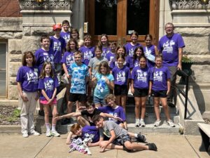 A group of children and adults pose on the steps of a building. They are wearing colorful t-shirts, mostly purple, with the same logo. Everyone seems happy. The building is made of stone, with a wooden door and decorative carvings.