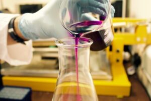 A gloved hand is pouring a purple liquid from a beaker into a conical flask. The background features laboratory equipment, including a yellow apparatus, suggesting a scientific setting.