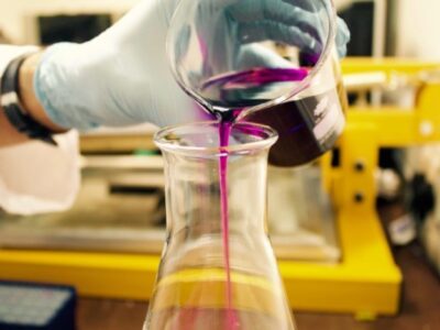 A gloved hand is pouring a purple liquid from a beaker into a conical flask. The background features laboratory equipment, including a yellow apparatus, suggesting a scientific setting.