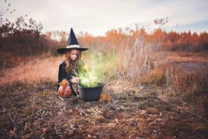 A child in a witch costume sits outdoors with a black cauldron emitting green smoke. A carved pumpkin decorates the scene. The background features dry autumn foliage under a cloudy sky.