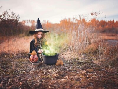 A child in a witch costume sits outdoors with a black cauldron emitting green smoke. A carved pumpkin decorates the scene. The background features dry autumn foliage under a cloudy sky.