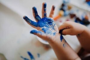 A child's hand covered in blue paint, with fingers spread open. The other hand is also partially visible, with paint smudges. Blurred background with assorted colors suggests a painting activity.