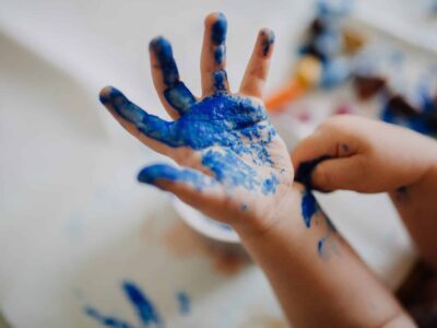 A child's hand covered in blue paint, with fingers spread open. The other hand is also partially visible, with paint smudges. Blurred background with assorted colors suggests a painting activity.