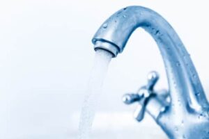 A close-up of a metallic faucet with water flowing steadily. The tap handle is designed with a cross shape, and droplets of water are visible on the faucet's surface. The background is a soft white, giving a clean and refreshing feel.