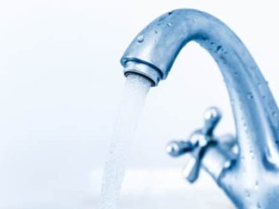 A close-up of a metallic faucet with water flowing steadily. The tap handle is designed with a cross shape, and droplets of water are visible on the faucet's surface. The background is a soft white, giving a clean and refreshing feel.