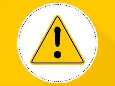 Yellow triangular warning sign with a black exclamation mark in the center, placed against a white circle with a dotted black border. The background is solid yellow, and the design casts a shadow to the right.