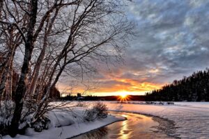 A winter landscape with a snow-covered riverbank and bare trees. The sun sets on the horizon, casting a warm glow over the snowy ground and partially frozen river. Dark clouds scatter across the sky, adding contrast to the orange hues of the sunset.