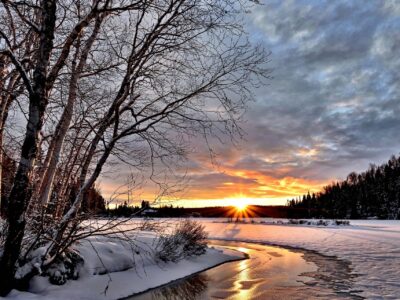 A winter landscape with a snow-covered riverbank and bare trees. The sun sets on the horizon, casting a warm glow over the snowy ground and partially frozen river. Dark clouds scatter across the sky, adding contrast to the orange hues of the sunset.