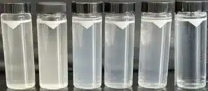 Six small, clear glass vials with black screw-on caps are lined up in a row. Each vial contains a translucent liquid, with slight variations in opacity visible among them.