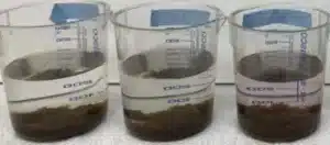 Three beakers filled with water and a sediment-like substance at the bottom are aligned next to each other on a surface. The beakers are transparent and have measurement markings and tape labels on them.