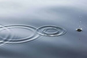 Water surface with rippling circles emanating from the point where a small droplet is splashing, creating a pattern of concentric waves against a smooth background.