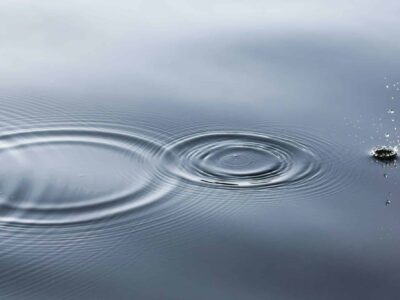 Water surface with rippling circles emanating from the point where a small droplet is splashing, creating a pattern of concentric waves against a smooth background.